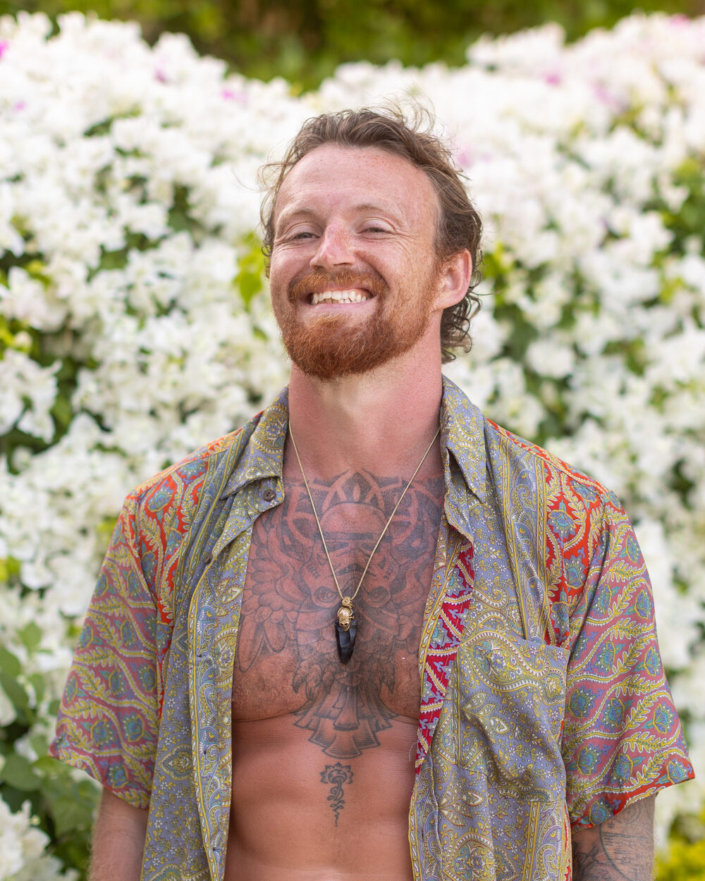 Ste Lane grinning in a paisley shirt in front of white bougainvillea
